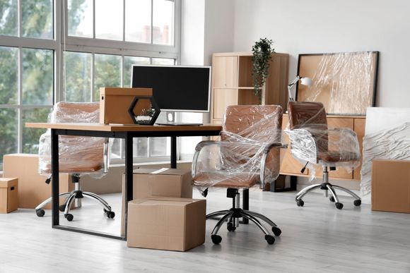 Empty office with wrapped desks, rolling chairs, cardboard boxes, and a monitor near large windows.