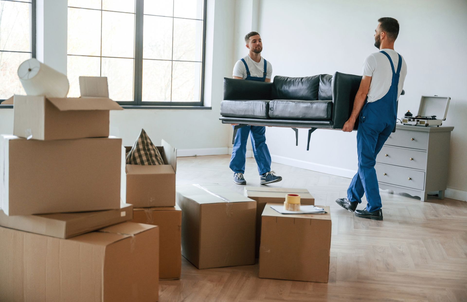 Two movers carrying a gray sofa through a bright room with stacked moving boxes