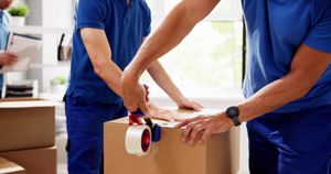 Movers in blue shirts sealing a cardboard box with packing tape in a bright room