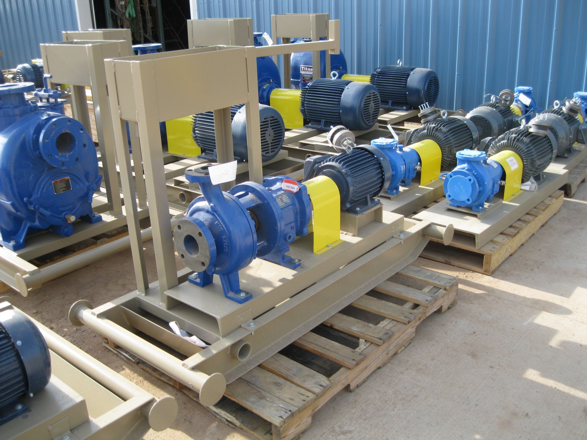Blue industrial pumps and motors mounted on wooden pallets outdoors beside a corrugated metal wall