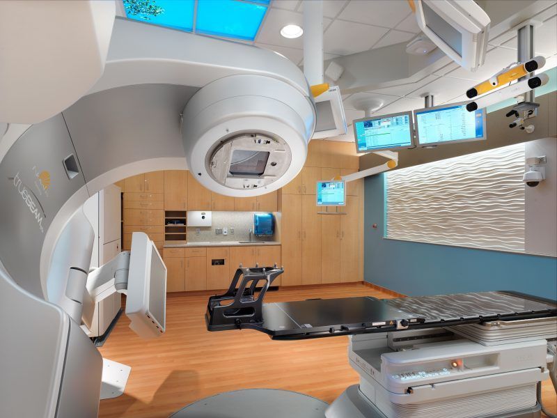 A linear accelerator in a modern, brightly lit clinical radiation therapy treatment room with a patient treatment table.