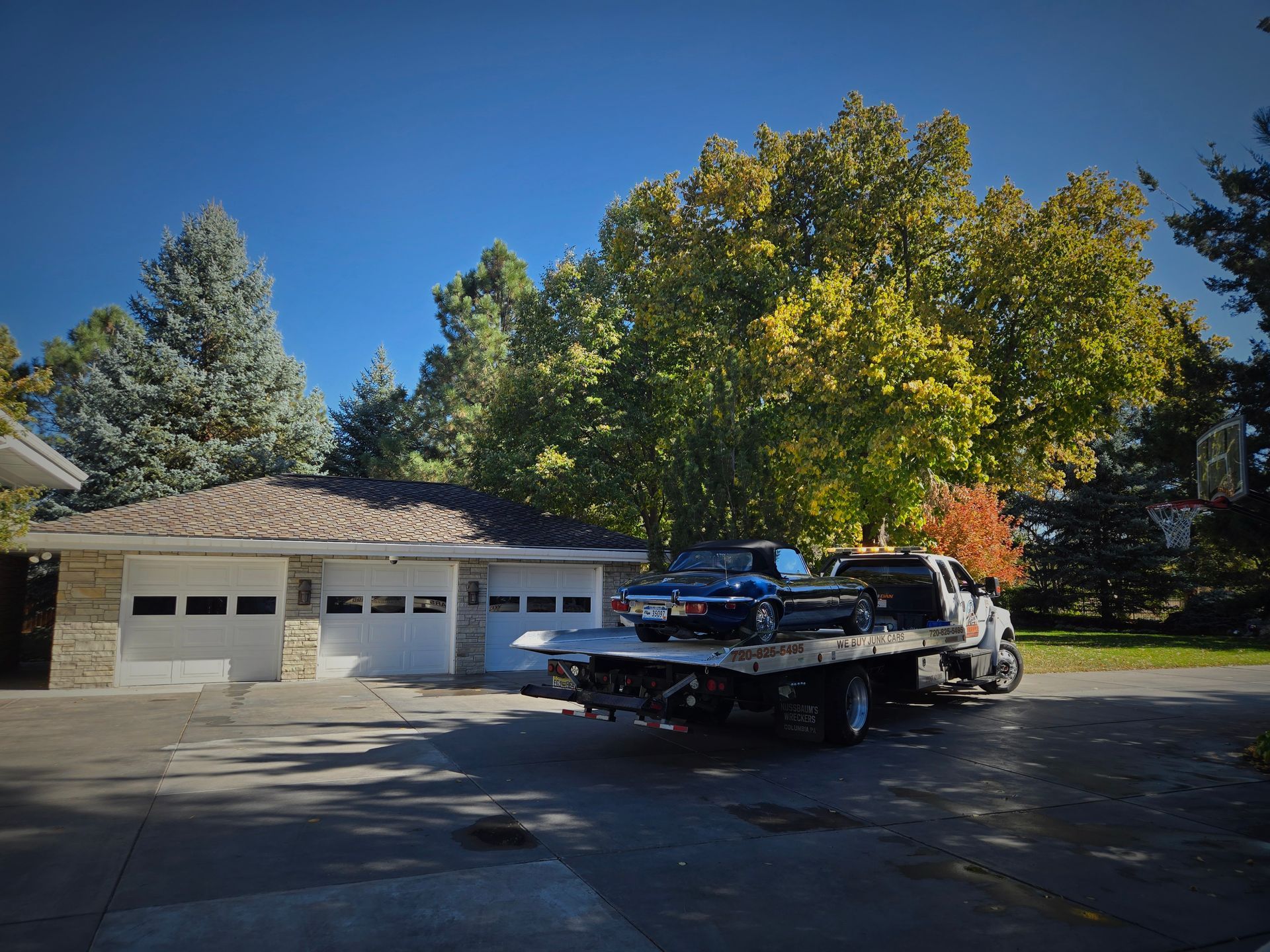 A dark car loaded onto a flatbed tow truck in a driveway in front of a three-car garage on a sunny day.