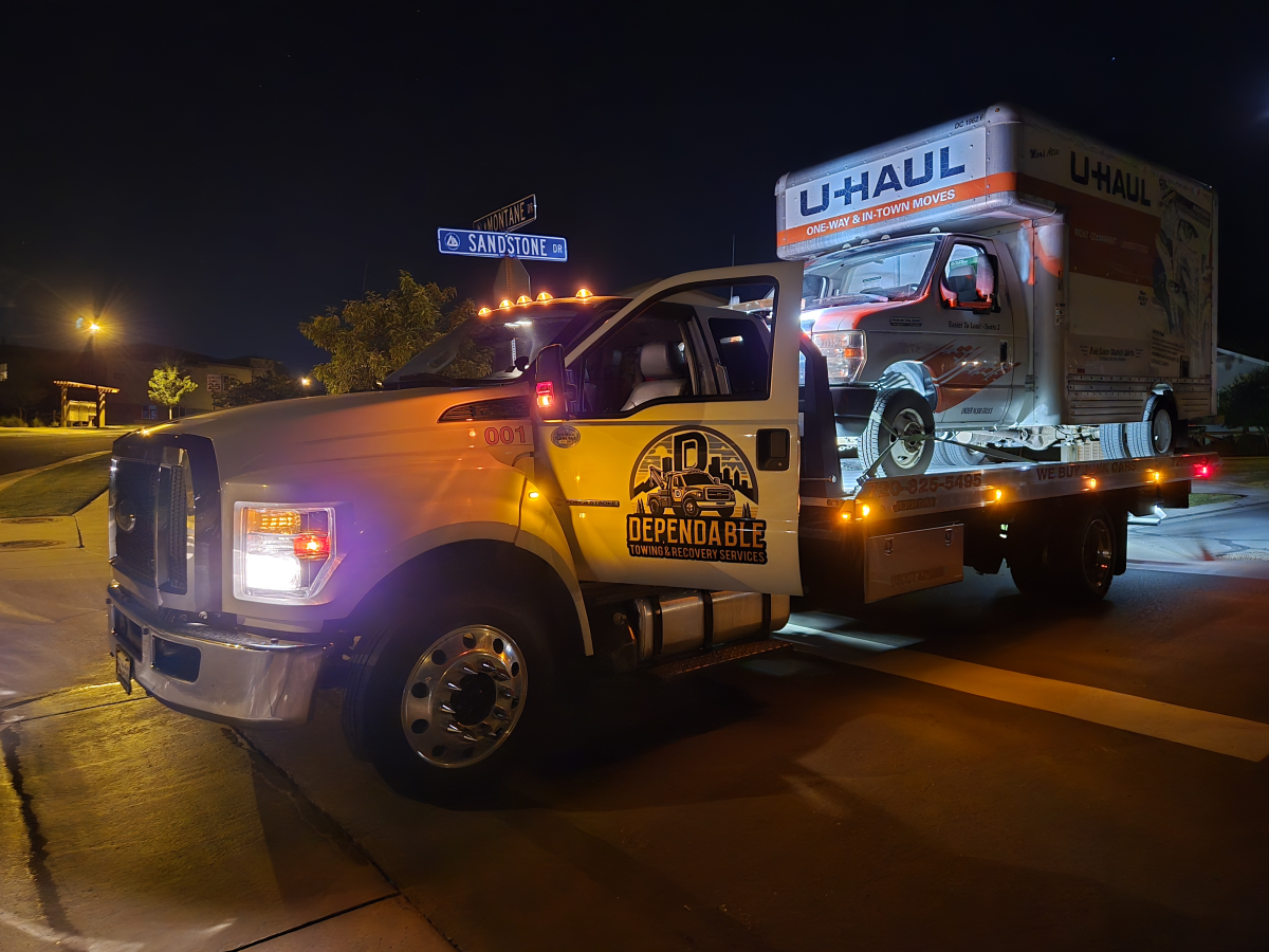 A white tow truck transports a U-Haul moving truck at night on a paved road.