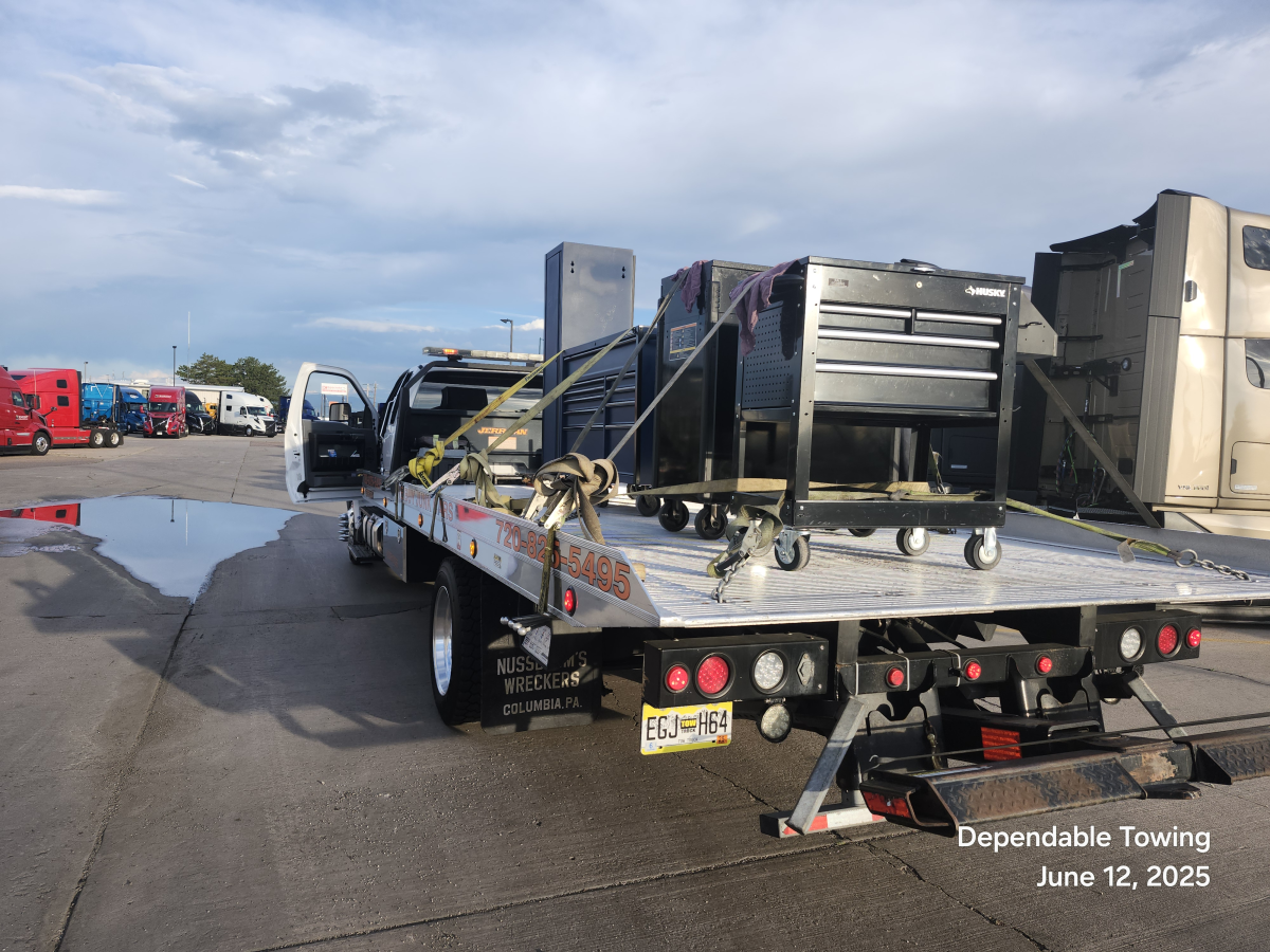 A flatbed tow truck parked in a lot carrying a large black rolling tool chest secured with straps.
