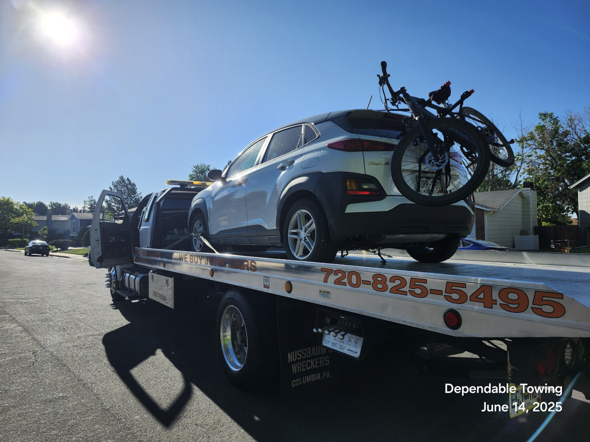 A white Hyundai Kona with two bicycles attached to the back is loaded onto a flatbed tow truck on a sunny day.