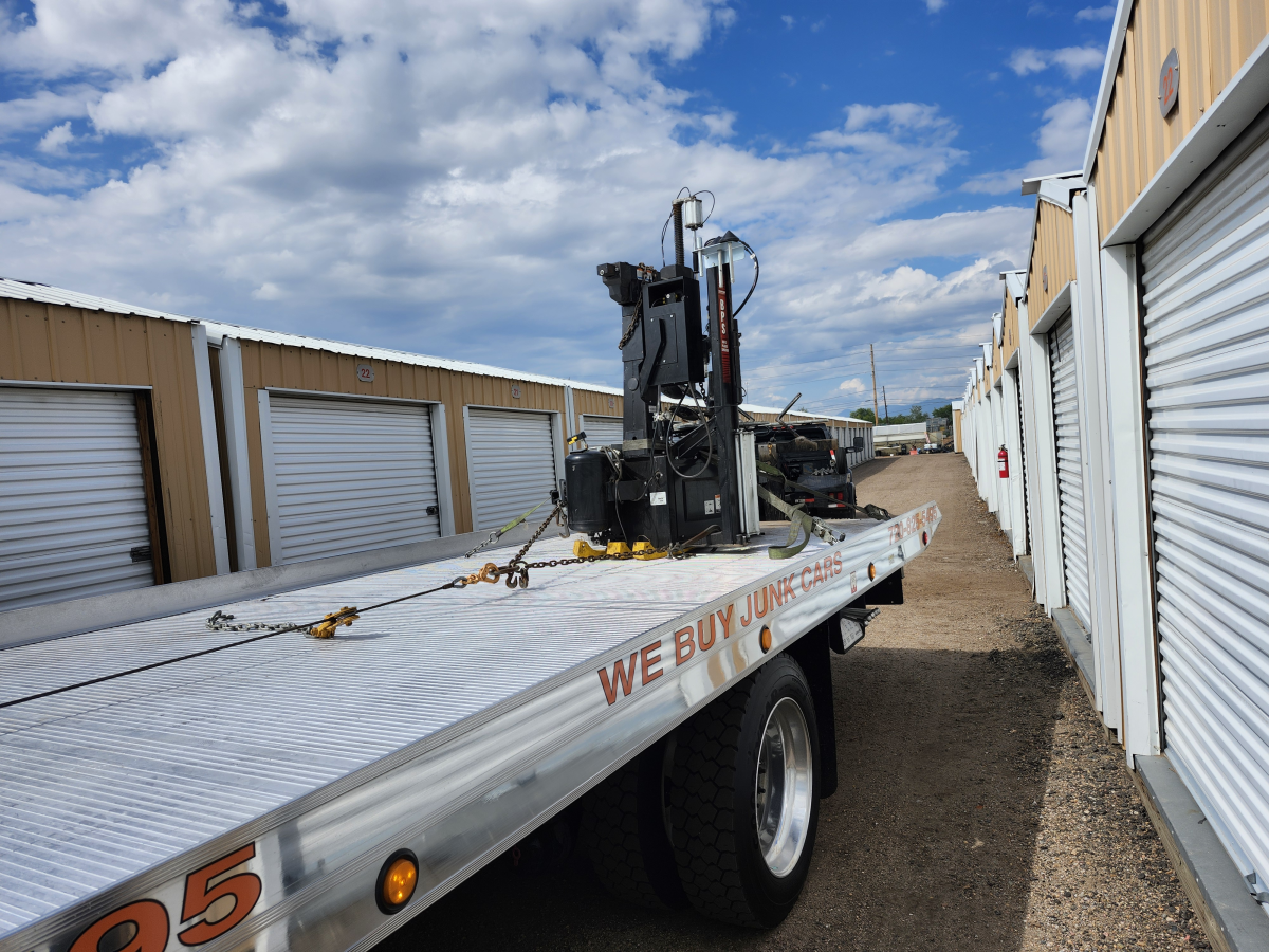 A flatbed tow truck parked between rows of storage units carrying a black piece of heavy machinery secured by chains.