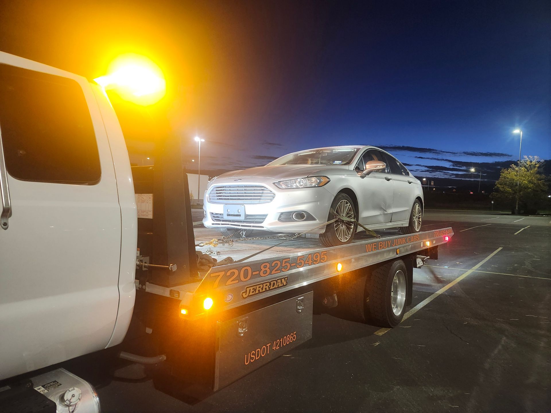 A silver Ford Fusion sedan sits secured on the flatbed of a tow truck at dusk, illuminated by glowing orange warning lights.