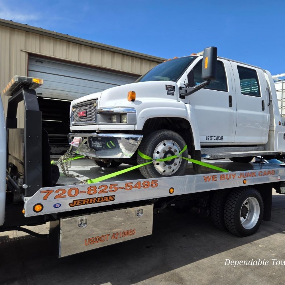 A white GMC truck is secured with green straps to the flatbed of a Jerr-Dan tow truck, parked in front of a tan building.