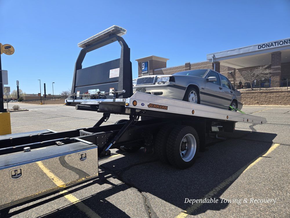A silver sedan loaded onto the tilted flatbed of a white tow truck in a parking lot on a sunny day.