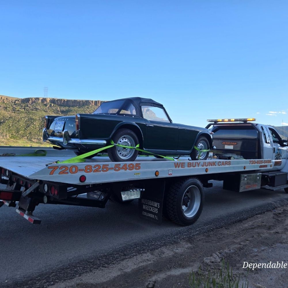 A dark vintage convertible sports car is secured to the flatbed of a white tow truck on a road against a hill background.