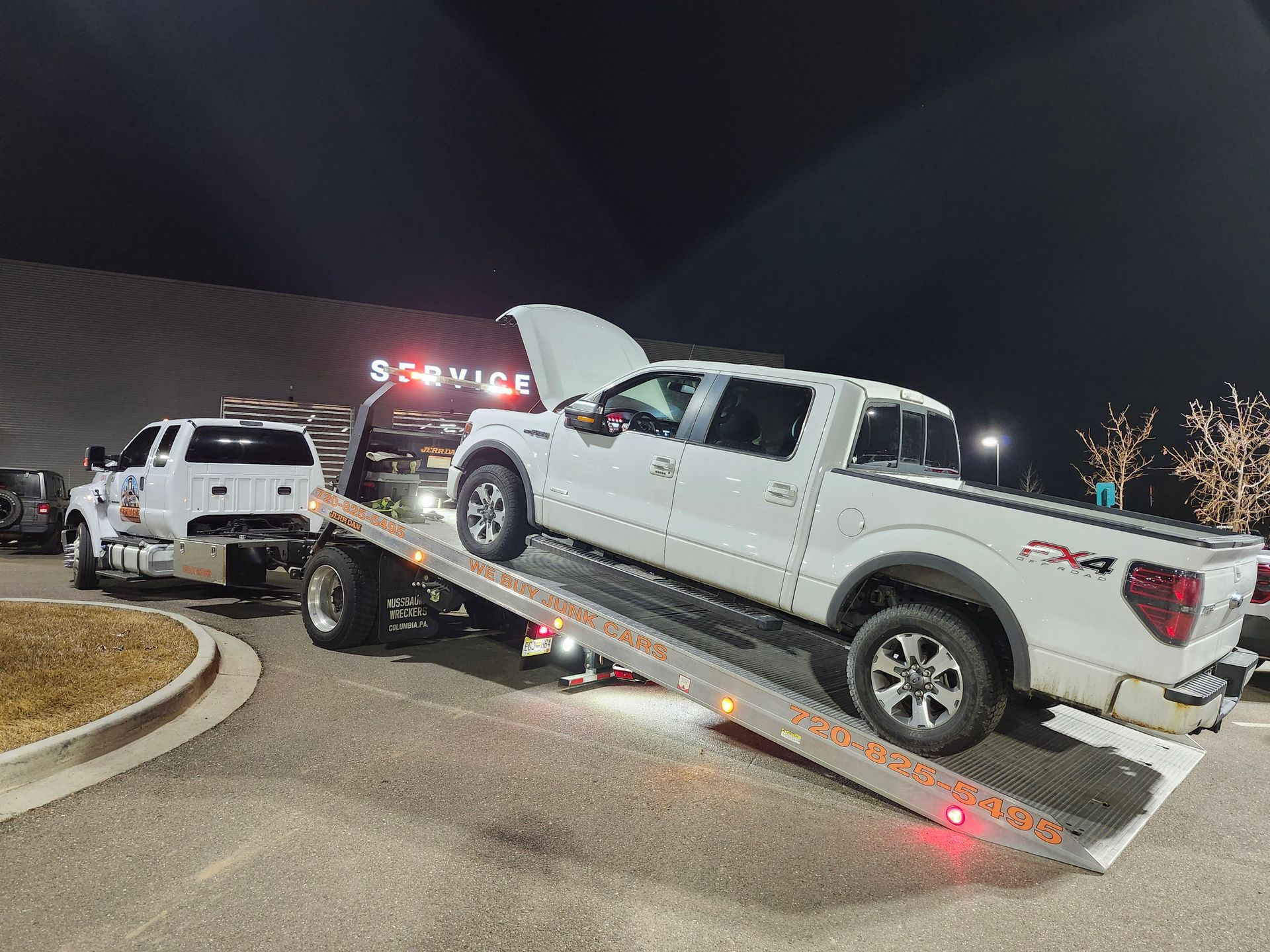 A white pickup truck with its hood open is being loaded onto a tow truck bed at night in a parking lot.