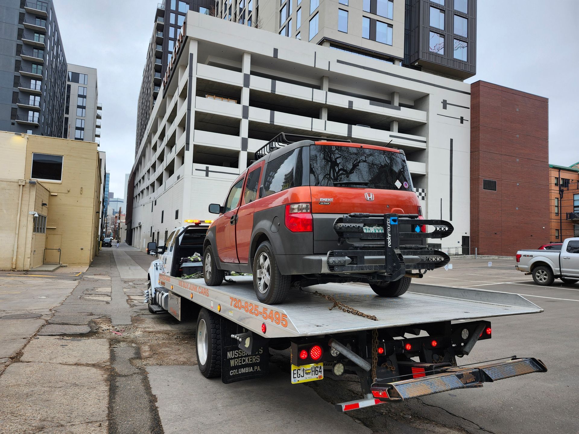 An orange Honda Element with a bike rack on the back is loaded onto the bed of a tow truck in an urban setting.
