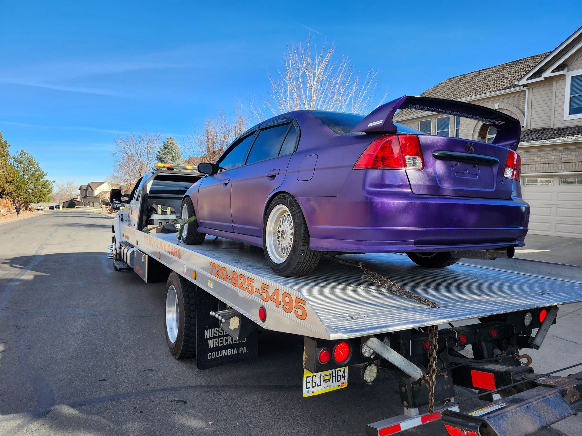 A purple sedan with a large rear spoiler is loaded onto the flatbed of a tow truck on a suburban street.