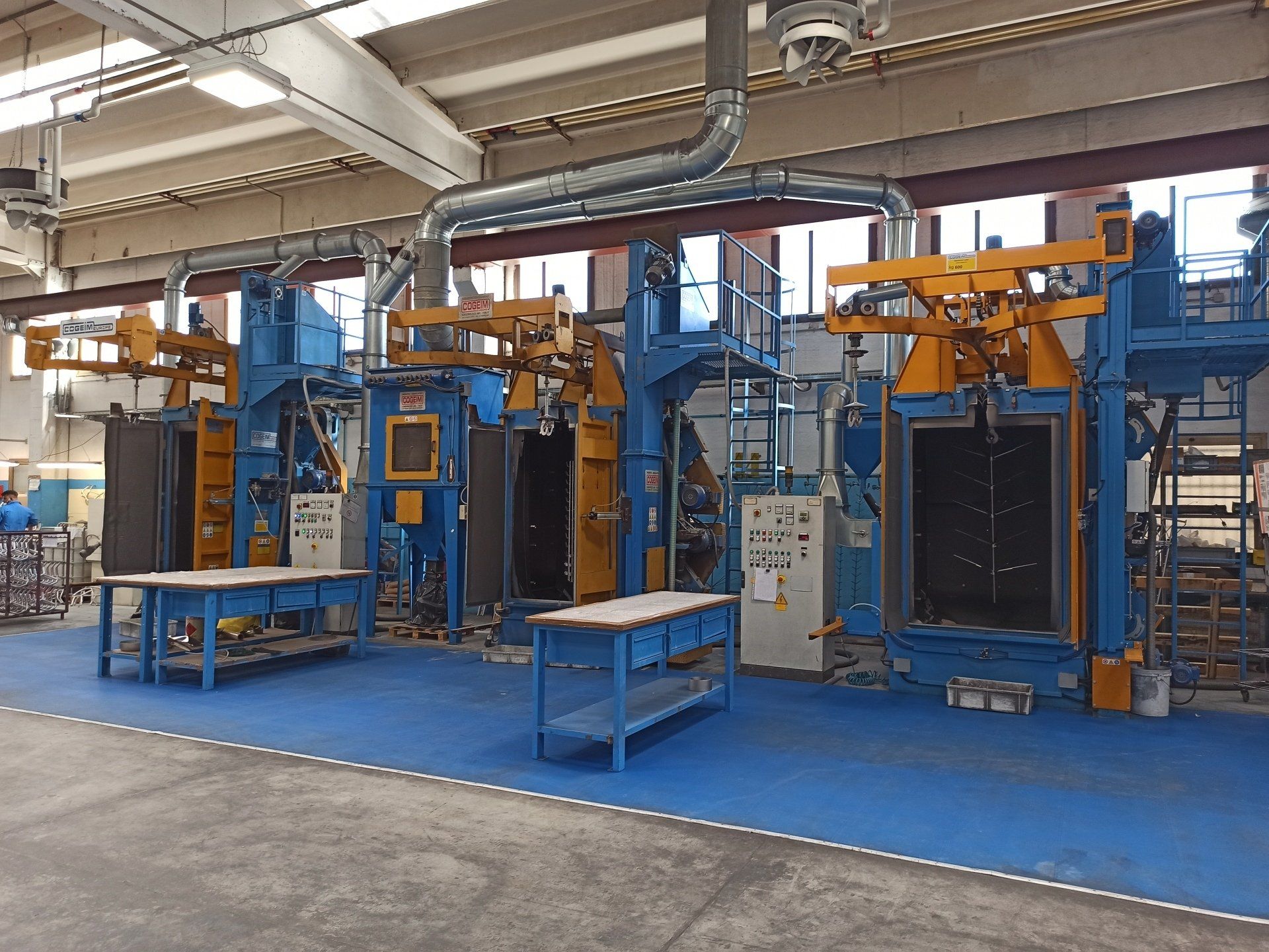 Tre macchine per la sabbiatura industriale in un ambiente di fabbrica, blu e gialle, con banchi da lavoro.