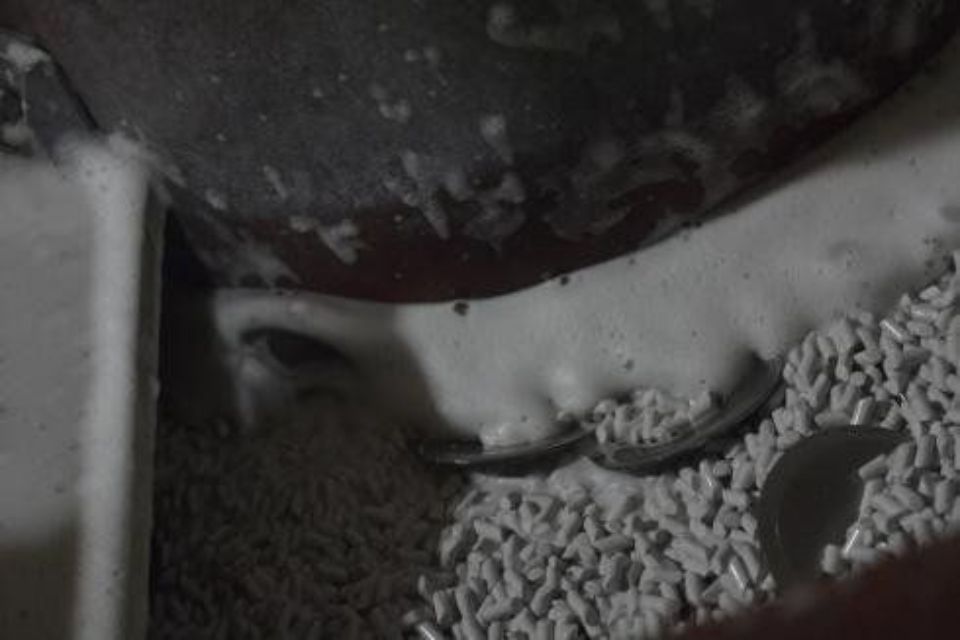 Primo piano di una macchina industriale che mescola pellet. La schiuma bianca trabocca, circondata da pellet di plastica grigia.