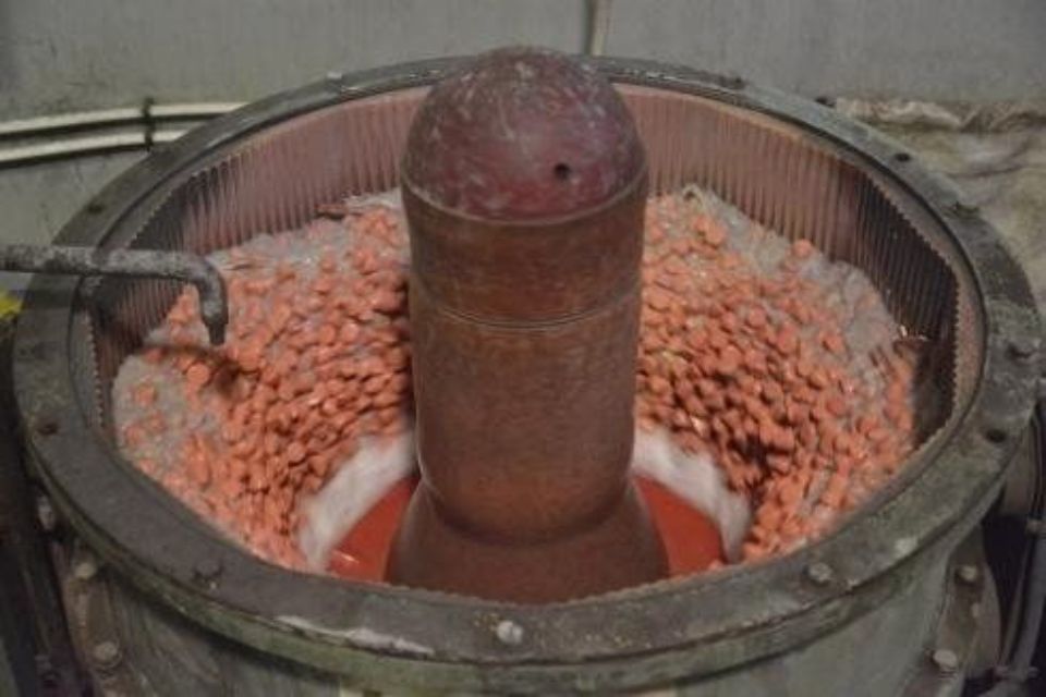 Un grande tamburo industriale, pieno di oggetti arancioni, agita un cilindro centrale in un ambiente industriale.