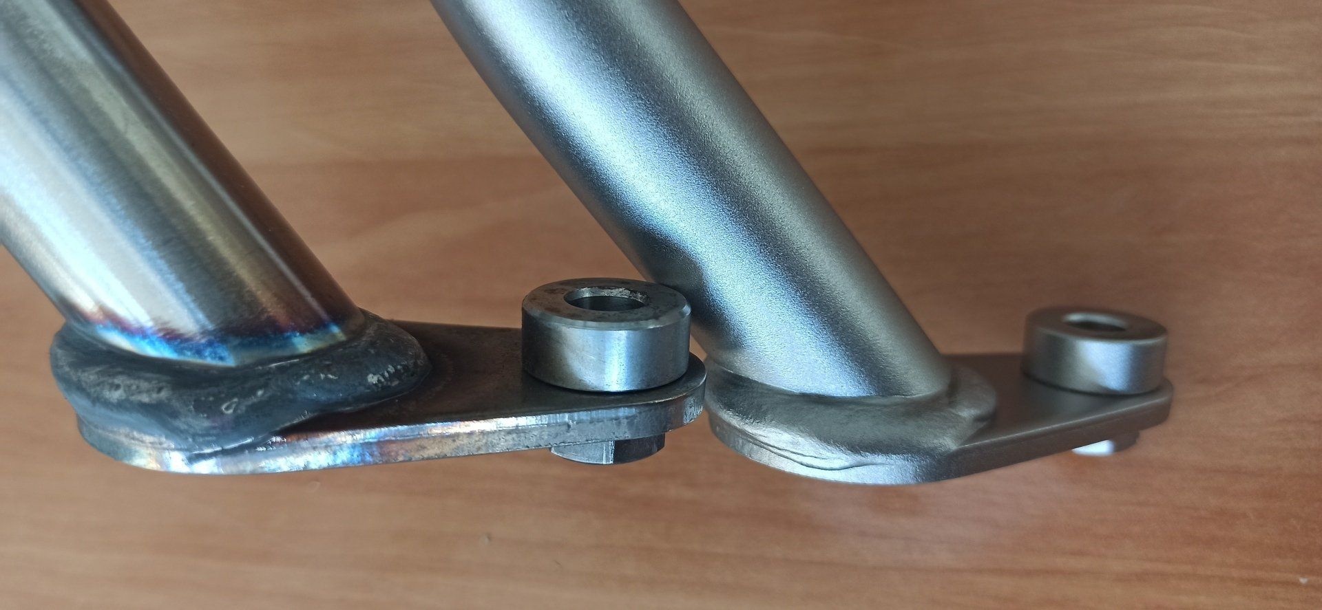 Due tubi metallici saldati a piastre con componenti cilindrici e piccole linguette, appoggiati su una superficie di legno.