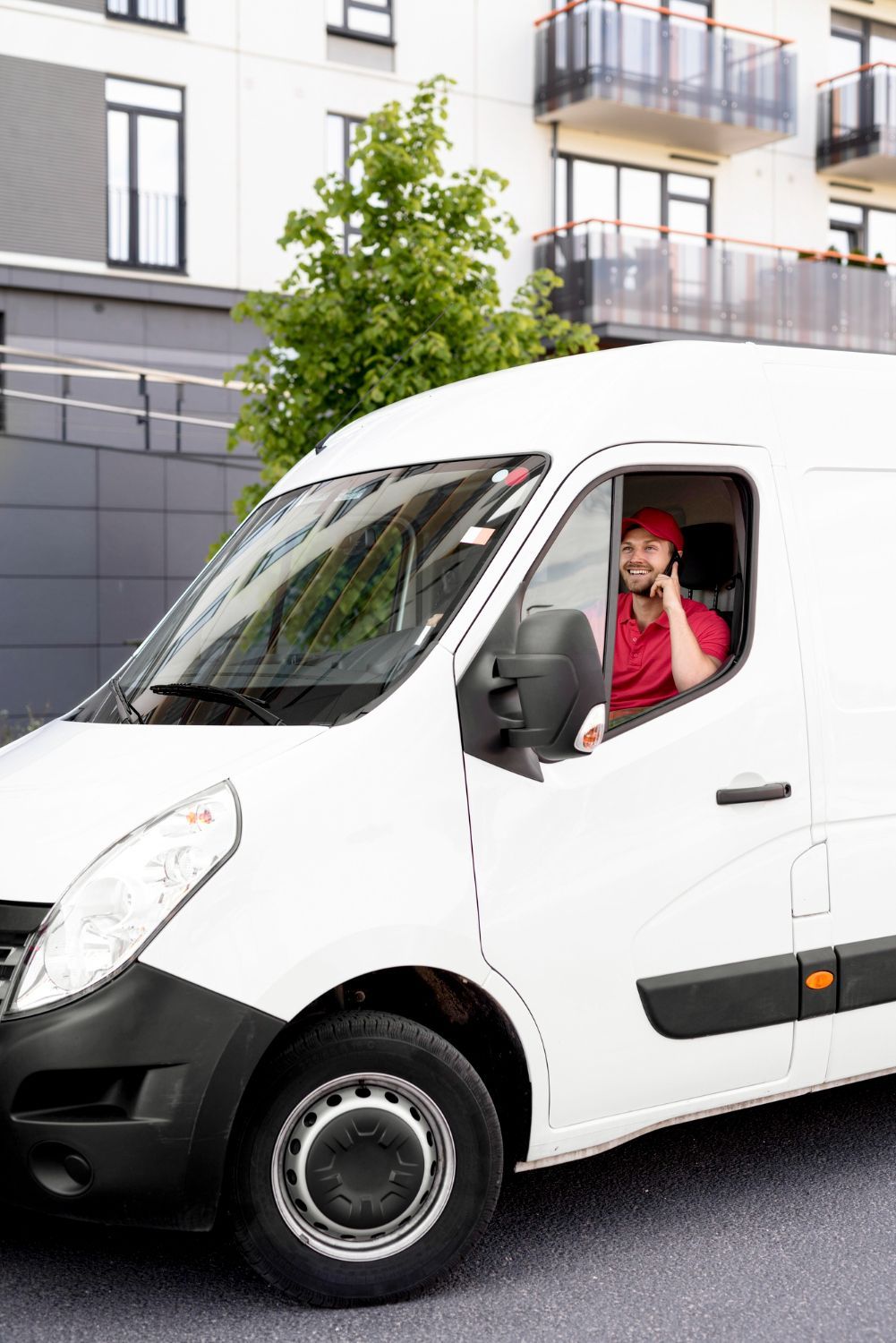 A delivery man is sitting in a white van talking on a cell phone.
