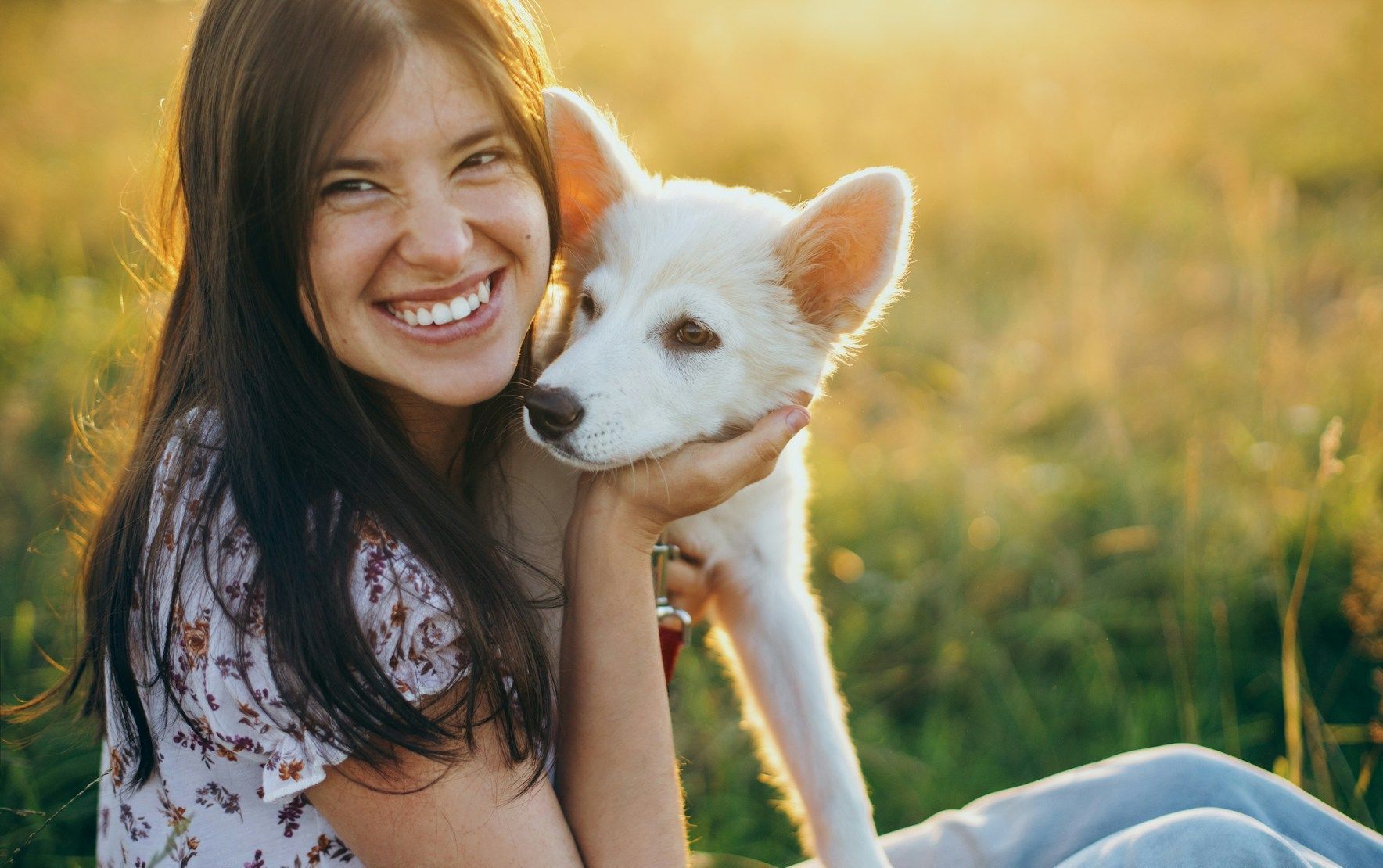A woman is sitting in the grass holding a white dog.