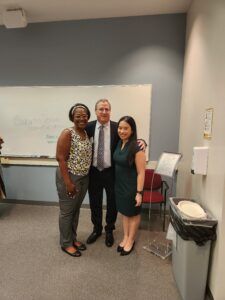 Three people pose in front of a whiteboard. A man in a suit stands between two women; they are smiling.