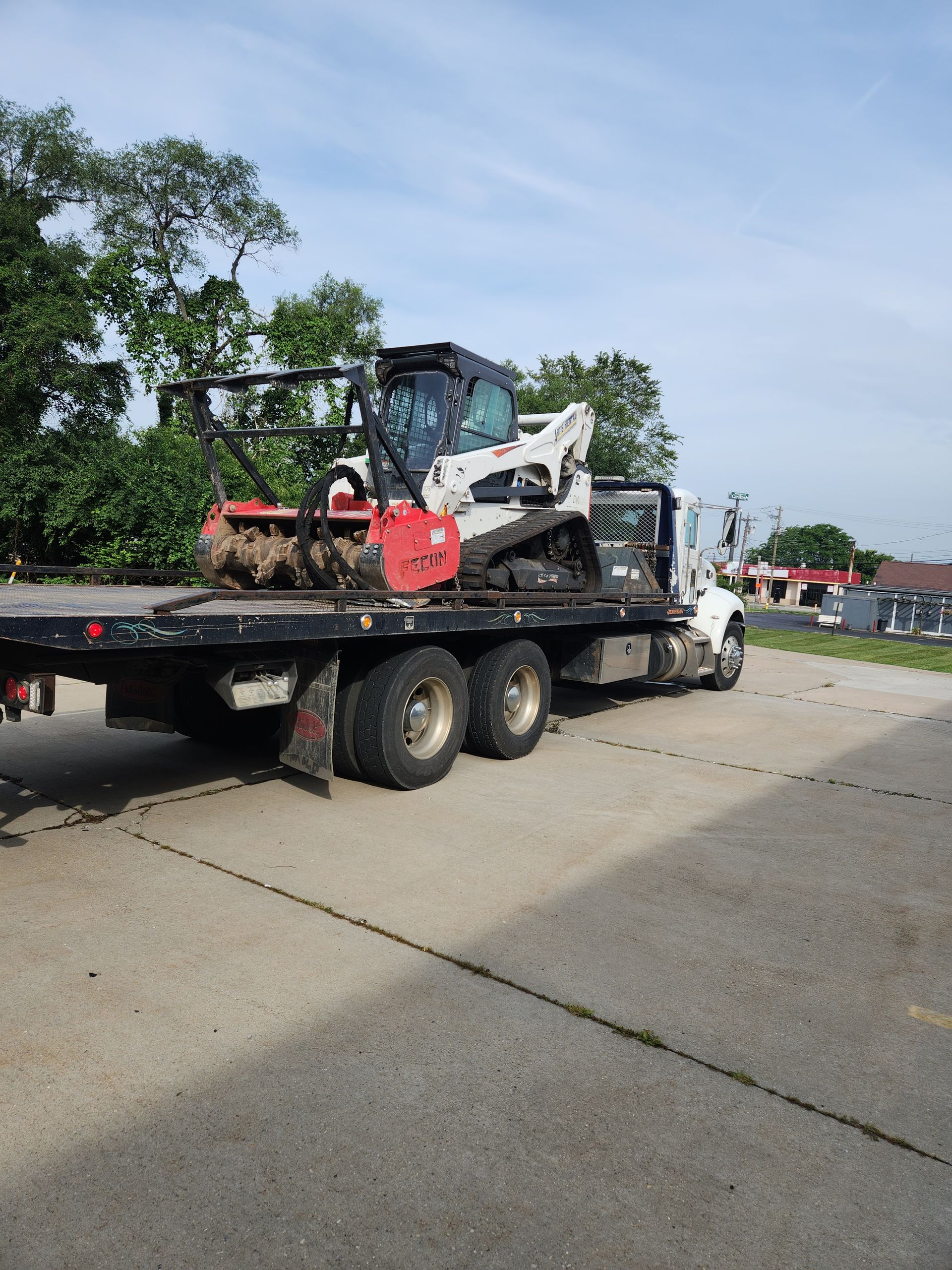 A white Bobcat compact track loader with a red mulcher attachment sits on a flatbed tow truck in an outdoor lot.