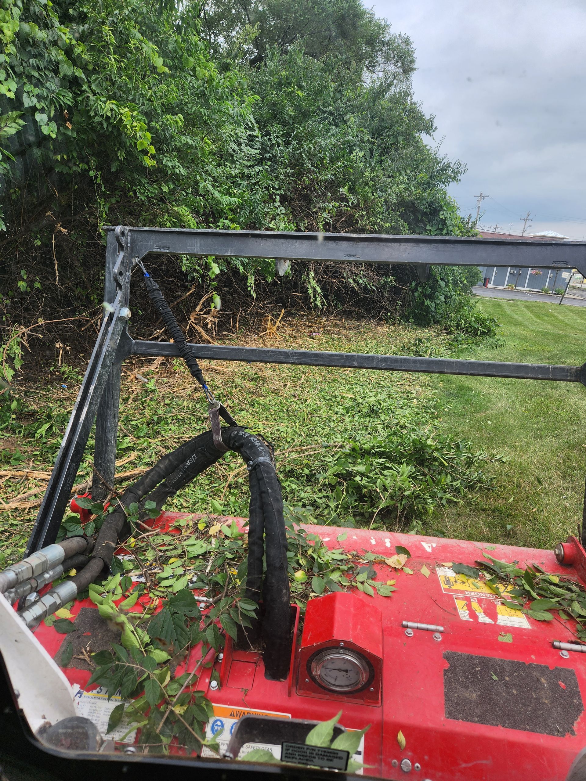 View from the cab of a red brush mower clearing dense green vegetation near a roadside.