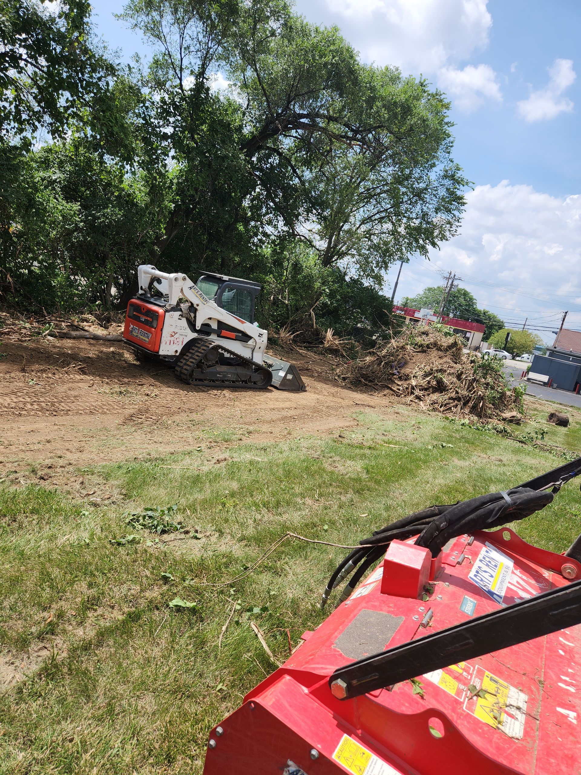 A white and red skid-steer loader clears brush in a grassy field with a red mulcher attachment in the foreground.