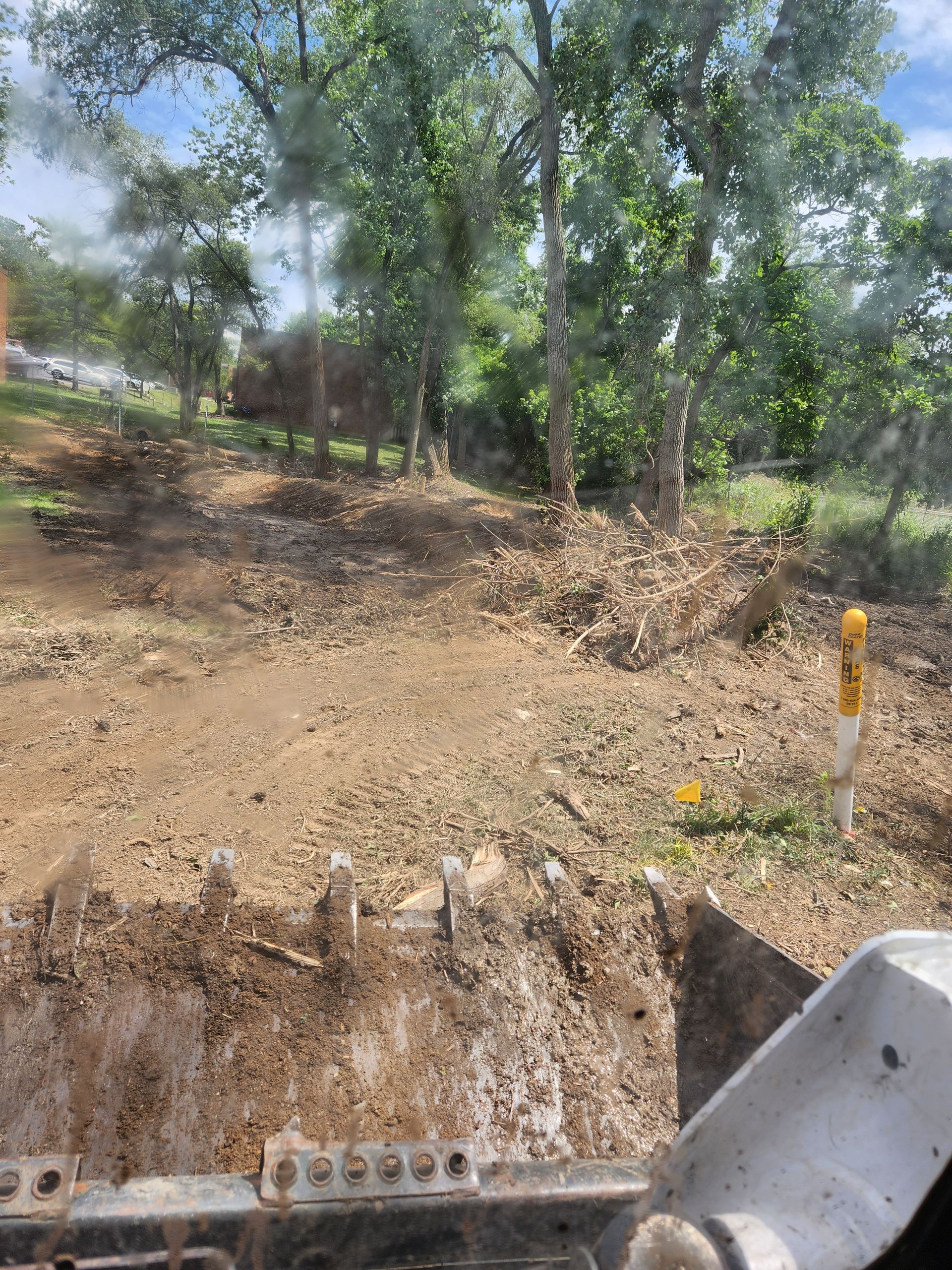 A view from an excavator cab looking over a cleared, wooded plot of land with a yellow utility marker post on the right.