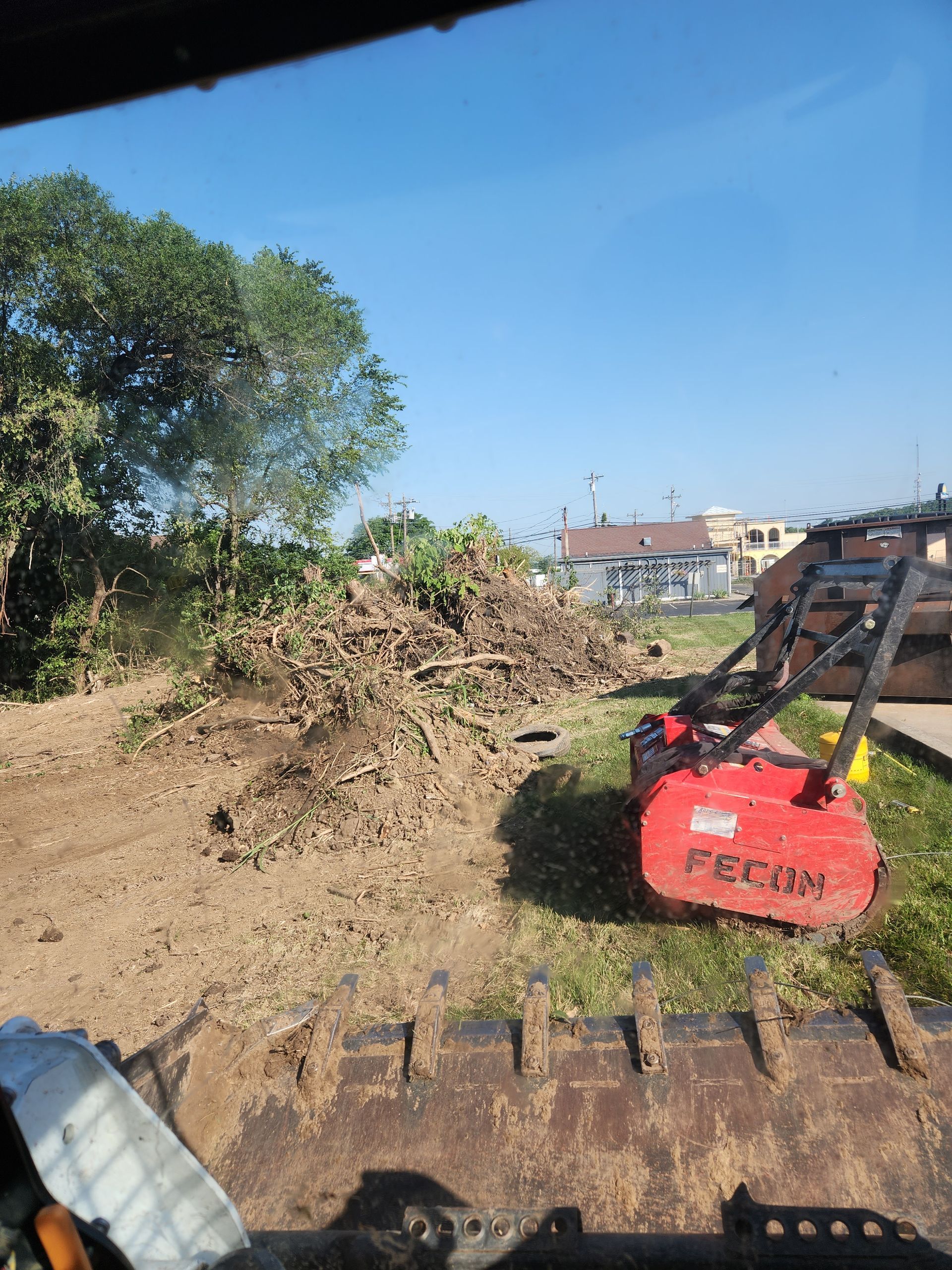 A red FAE mulcher attachment sits on a grassy field next to a large pile of brush and debris under a clear blue sky.