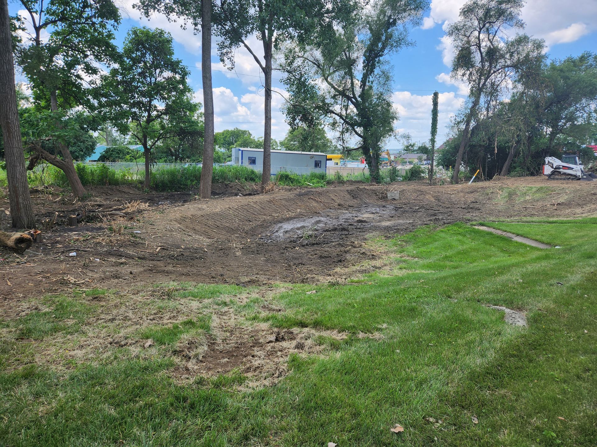 A dirt patch with tracks from heavy machinery bordered by green grass and trees under a blue sky with white clouds.