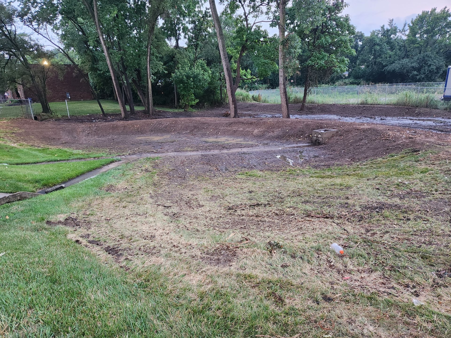 A view of a muddy, partially landscaped backyard with patches of green grass and a row of trees in the background.