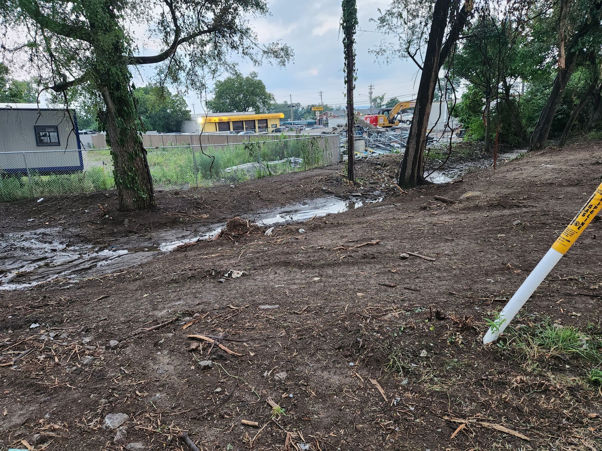 A muddy clearing with a small puddle, trees, a trailer on the left, and a distant building under a cloudy sky.