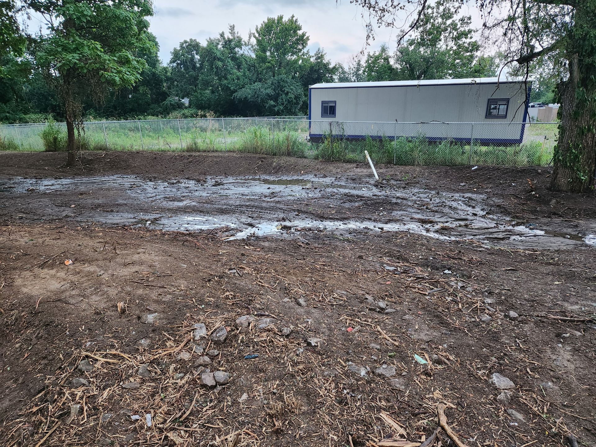 A muddy field with standing water in the foreground, near a white trailer and trees under a cloudy sky.