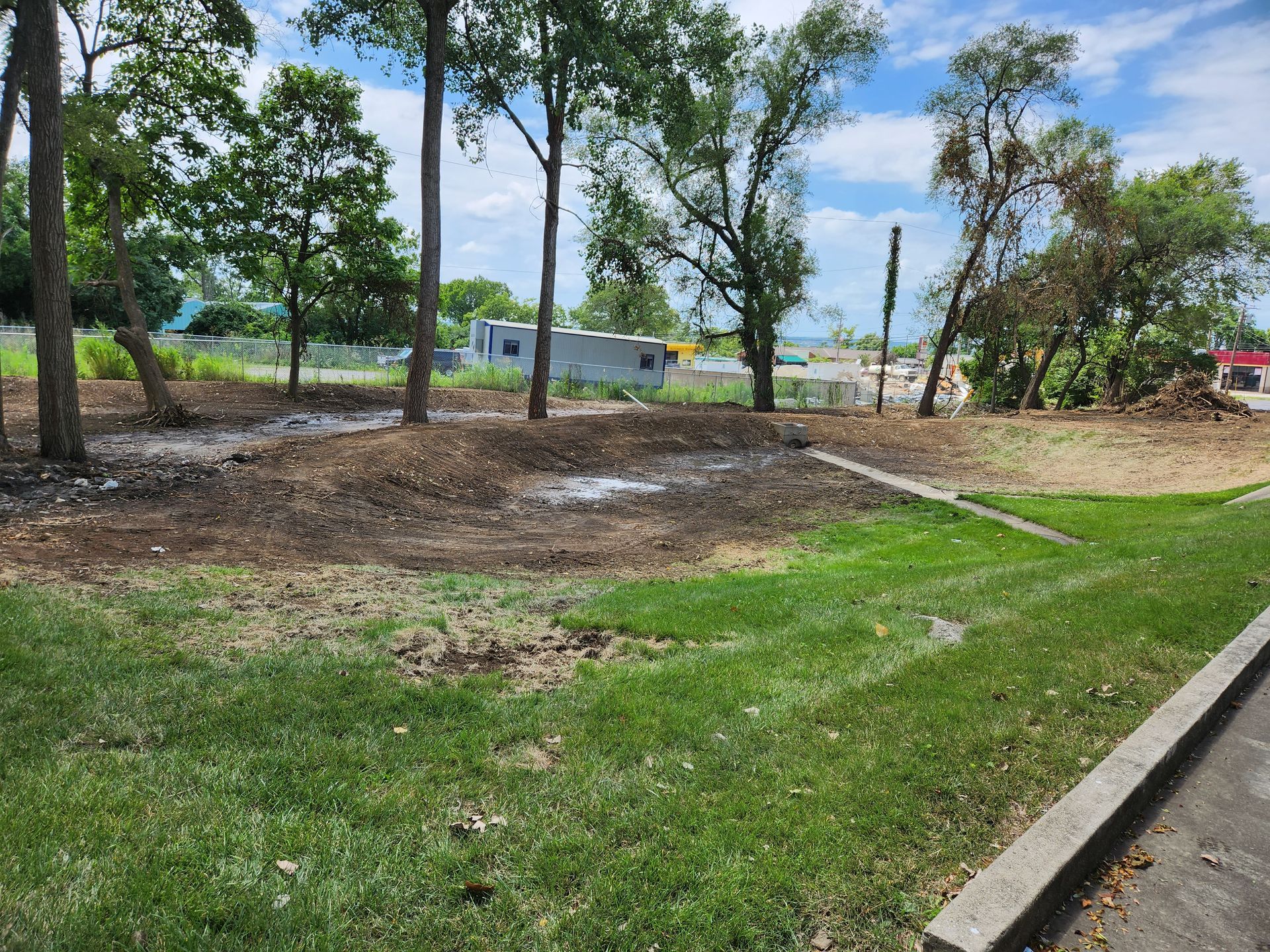 A grassy foreground leads to a cleared dirt lot with several trees, under a blue sky with clouds in the distance.