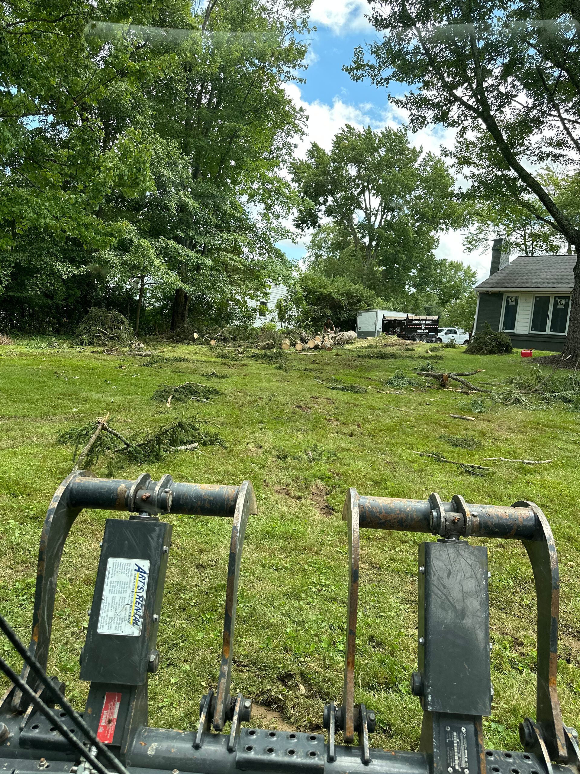A view from inside a tractor cab looking out at a grassy yard with trimmed brush, trees, and a house in the background.