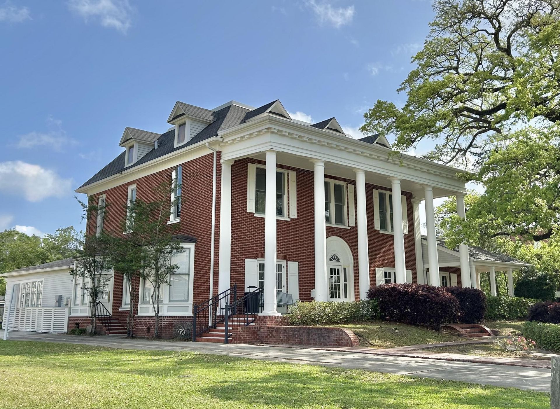 Historic red-brick house with white columns and wraparound porch on a grassy lawn under a blue sky