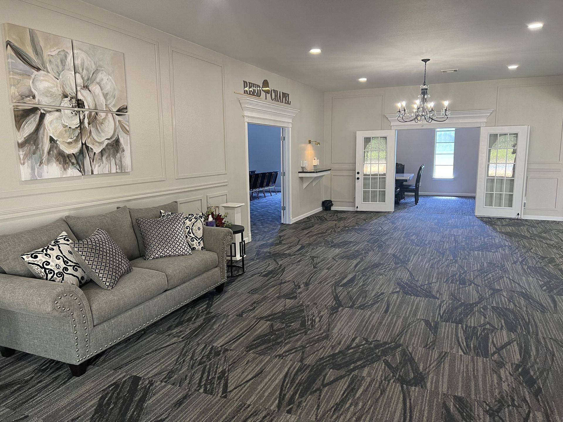 Bright lobby with gray sofa, floral wall art, chandeliers, and patterned carpet leading to doors.