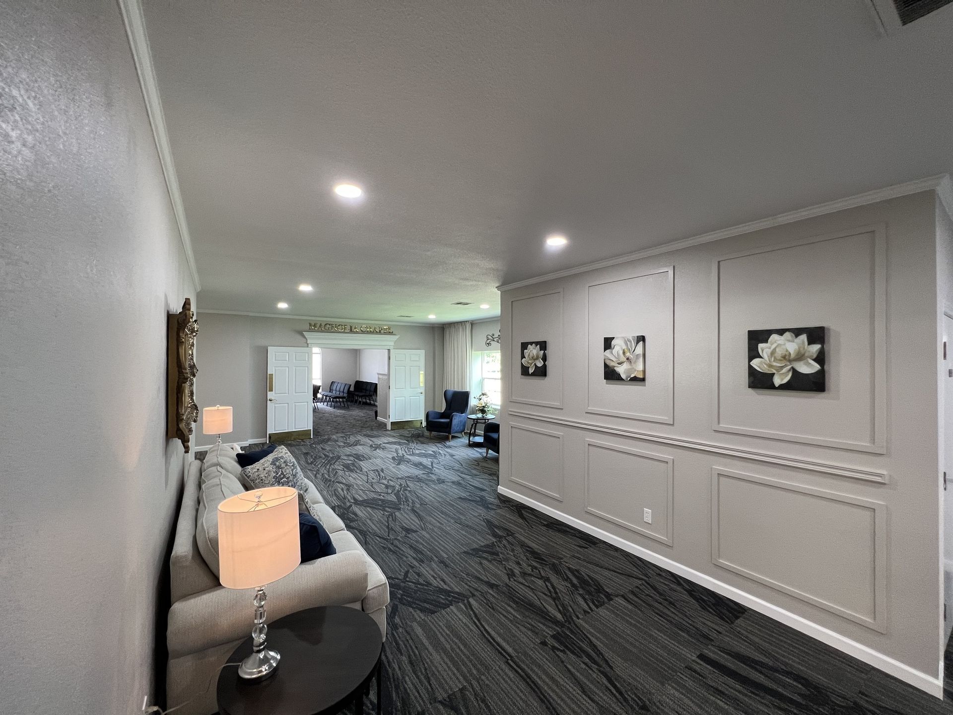 Modern hotel hallway with gray carpet, white walls, framed art, and a small seating area on the left