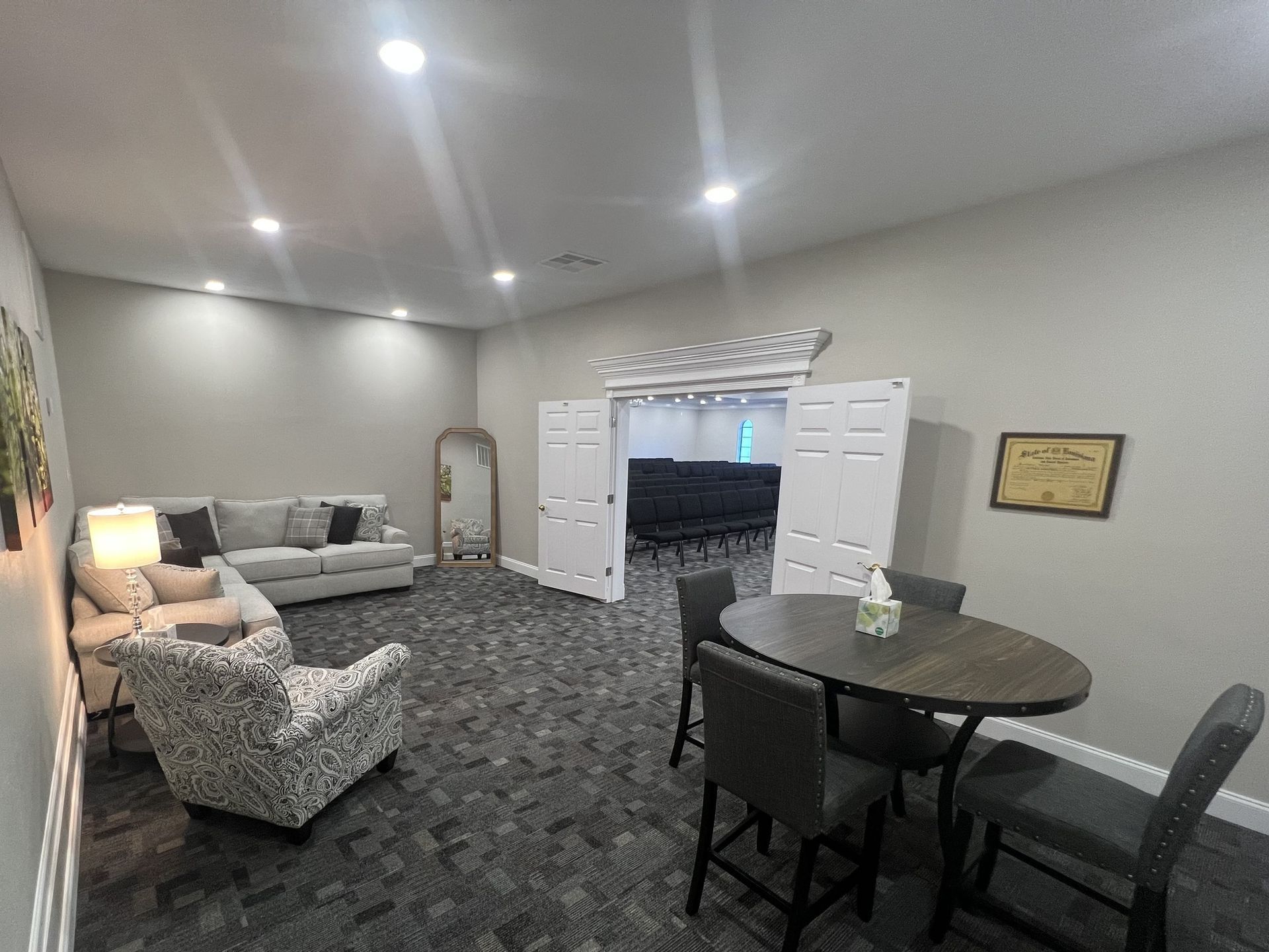 Bright living room with gray sofa, patterned armchair, round table, and double doors to a TV room