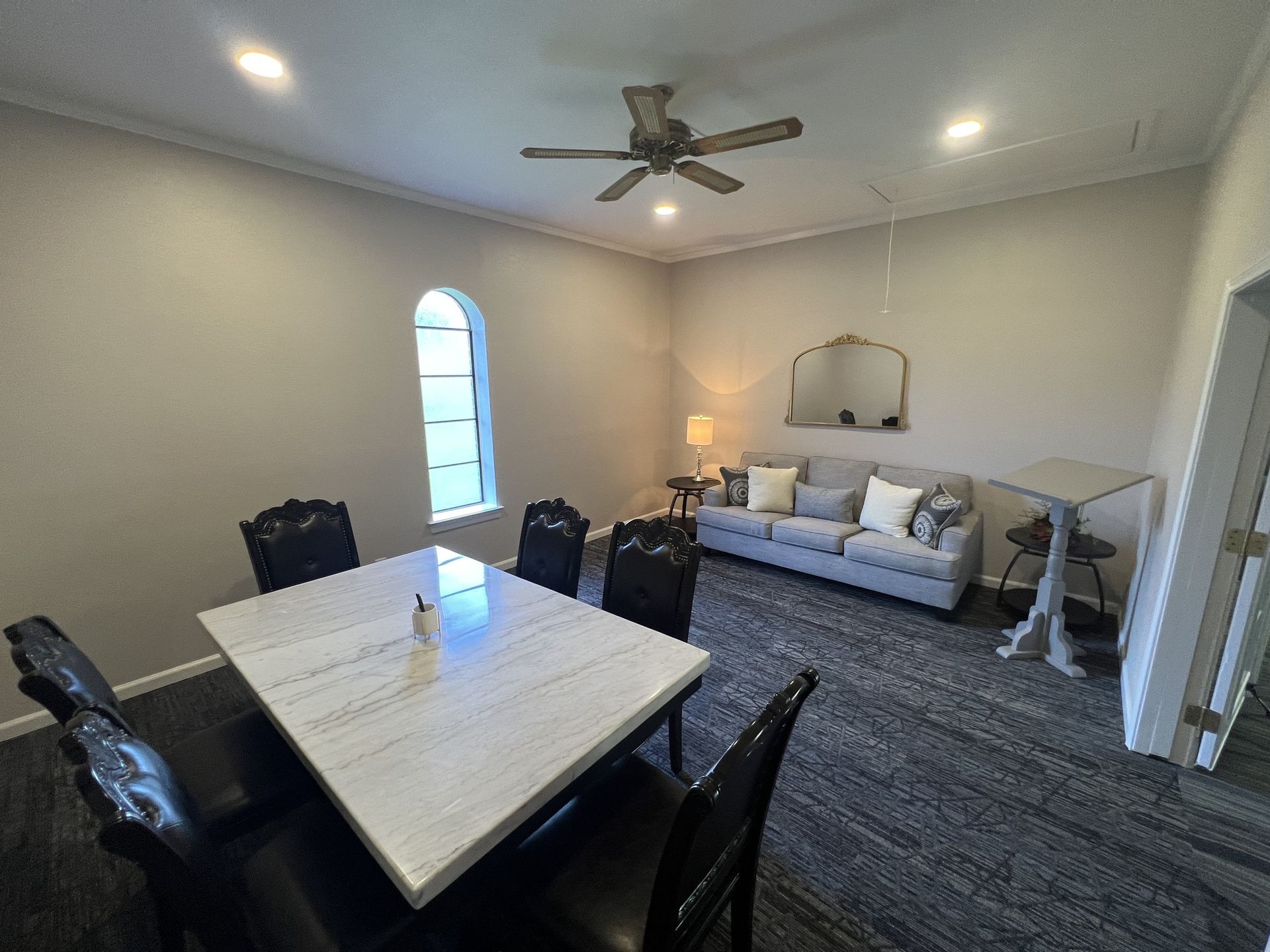 Living room with dining table, black chairs, gray carpet, couch, ceiling fan, and arched window.