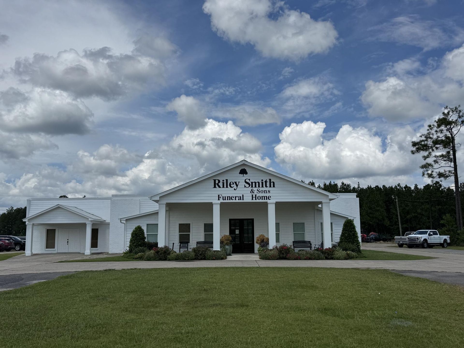 White church building with columns and a sign, set beside a grassy field under a cloudy sky.