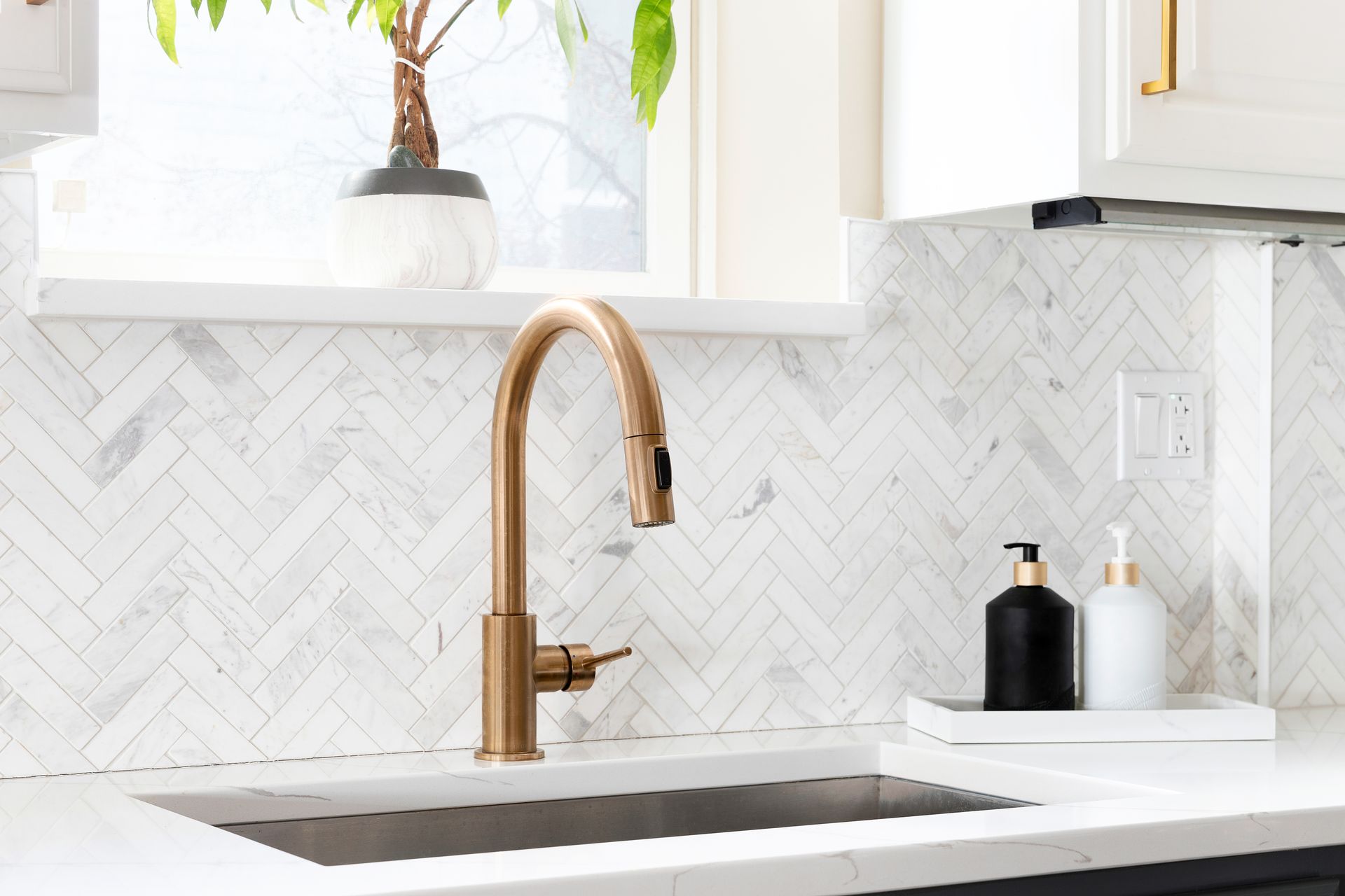 Bright kitchen sink with gold faucet, white herringbone backsplash, and soap bottles on a tray