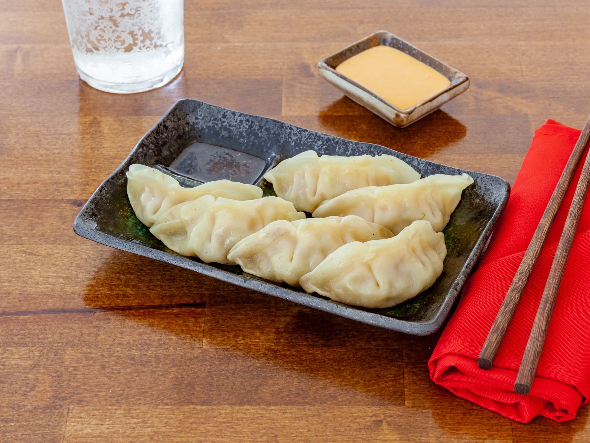 Plate of steamed dumplings on a wooden table with red chopsticks, sauce, and a glass of water
