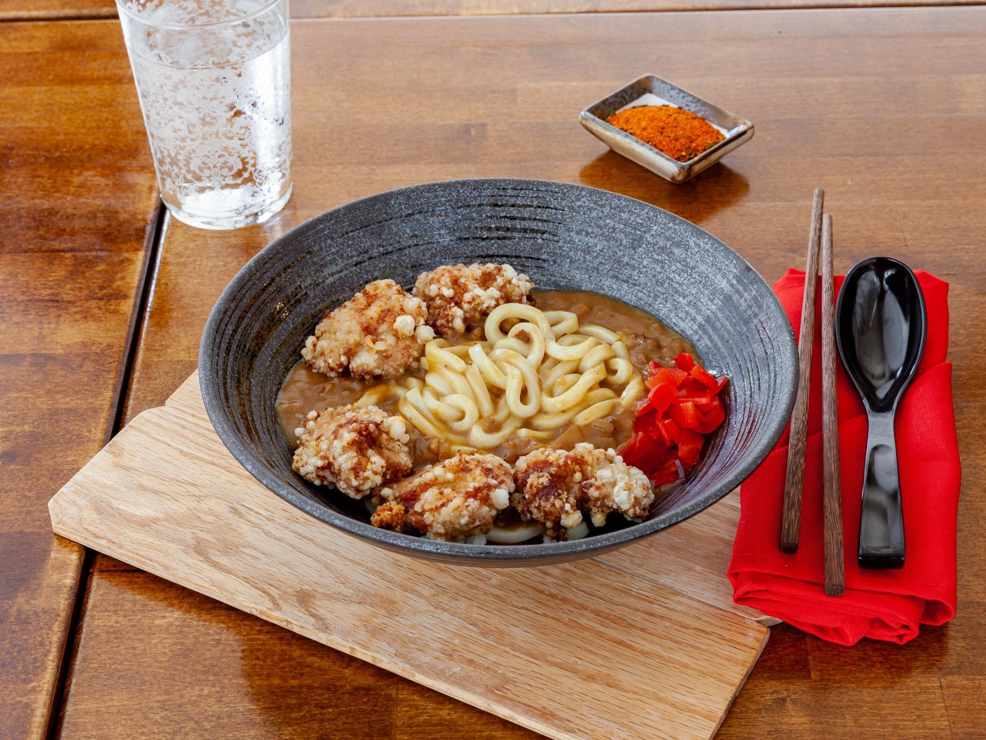 Bowl of udon noodles with meat on a wooden tray, with chopsticks, spoon, and water glass beside it.