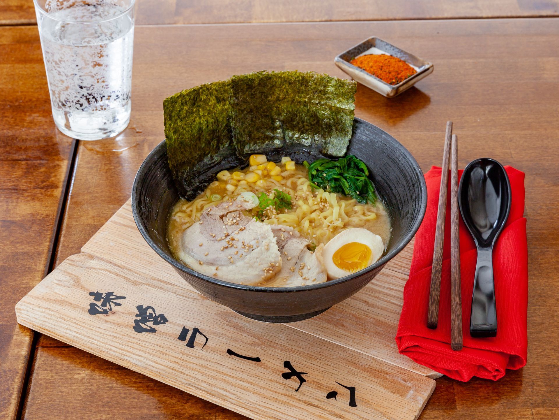 Ramen bowl with seaweed, egg, and greens on a tray beside chopsticks and a glass of water