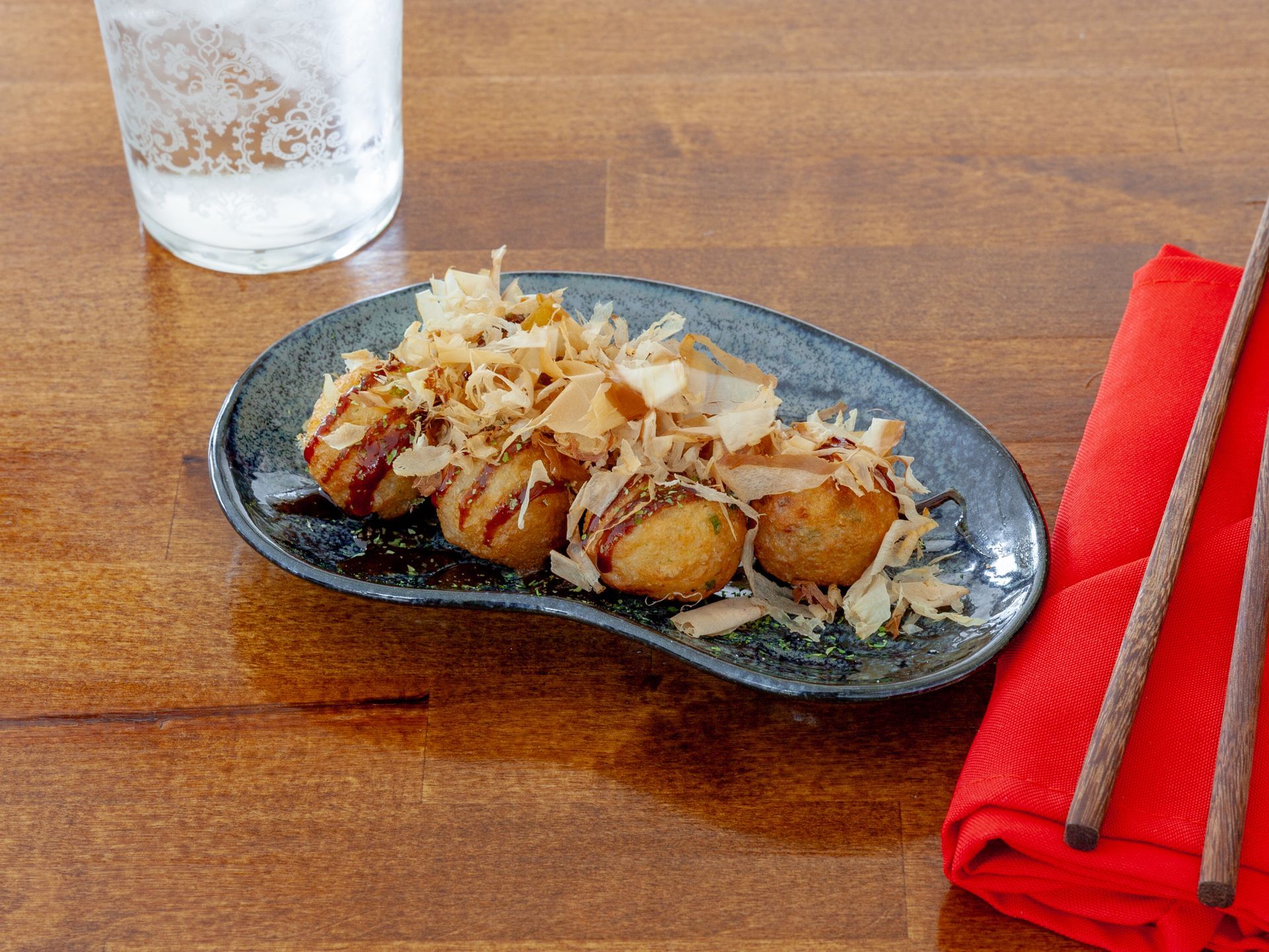 Grilled scallops topped with crispy garnish on a black plate beside a red napkin and chopsticks on a wooden table