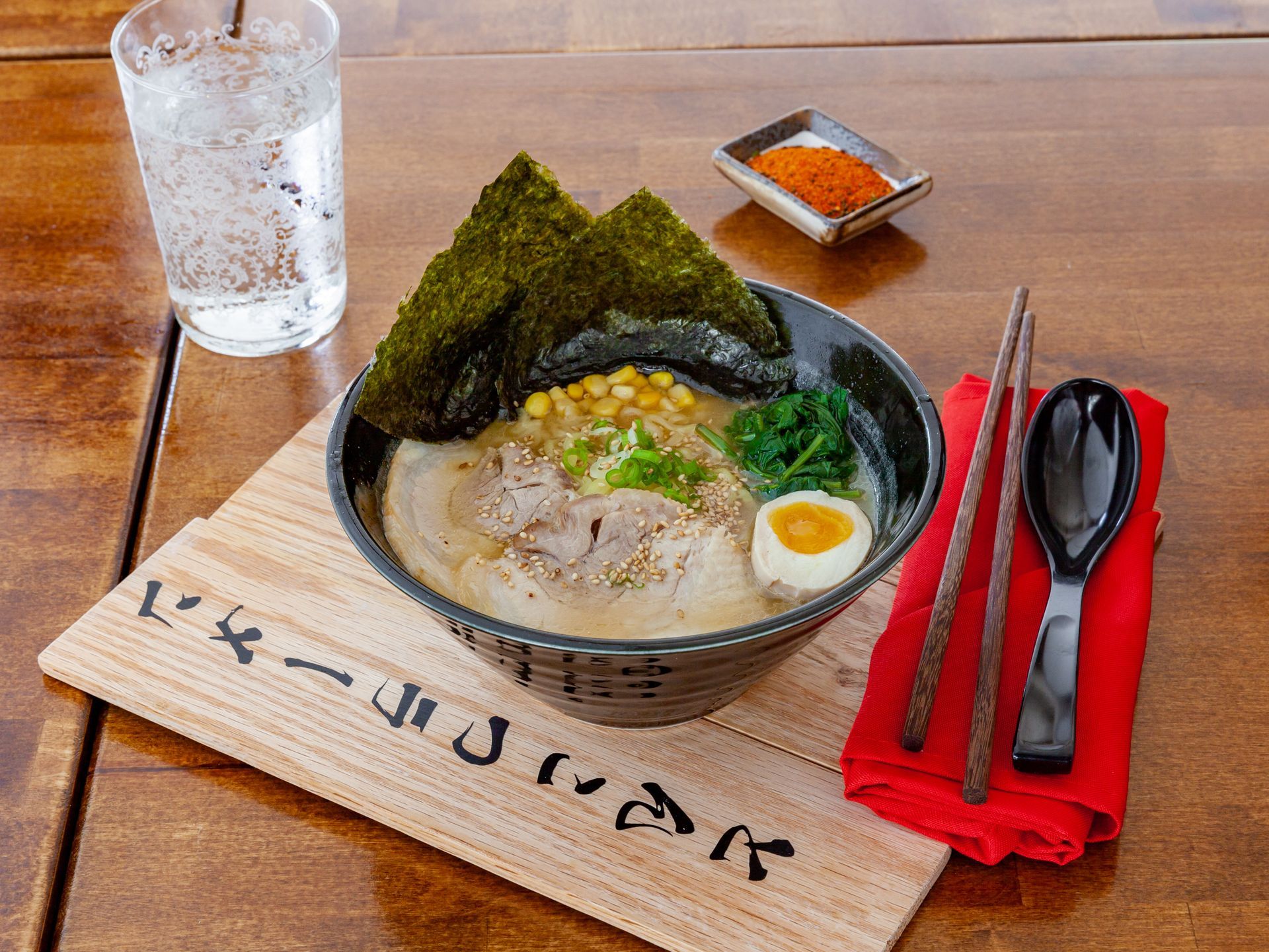 Ramen bowl with seaweed and egg, chopsticks, spoon, glass of water, and chili paste on a wooden table