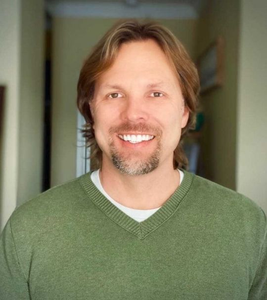 A man with a beard is wearing a green sweater and smiling.