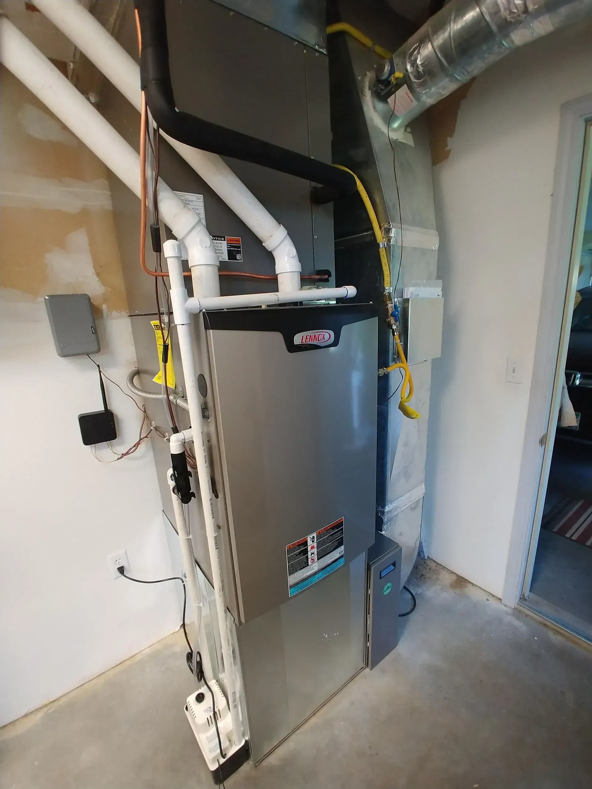 A furnace unit in a basement, with connected pipes and ducts. Grey and white, in a corner.