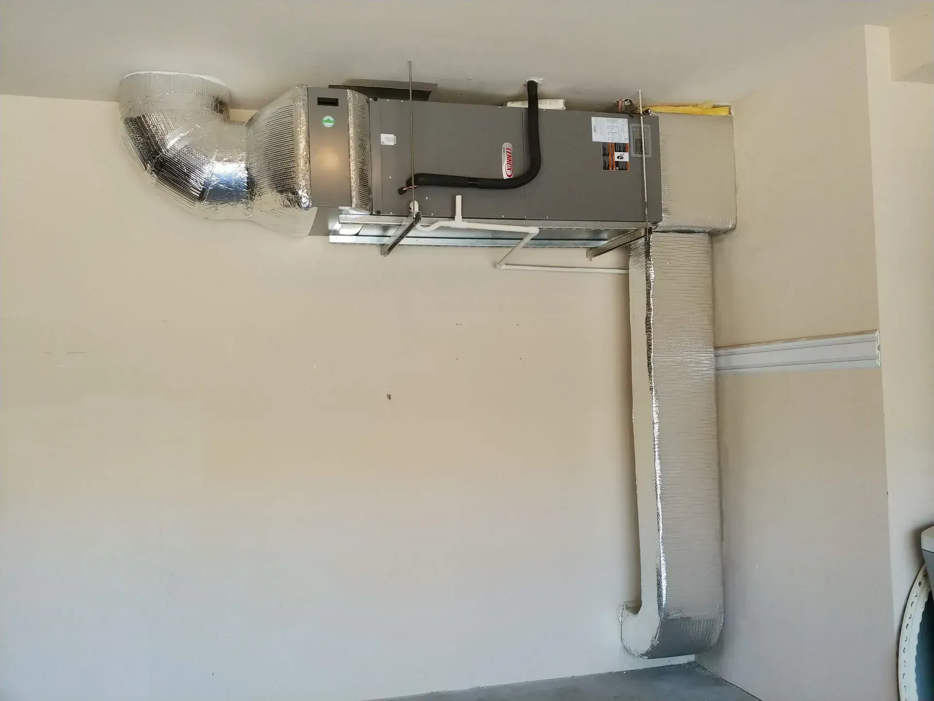 HVAC unit mounted on a beige wall in a room. Ductwork, silver-colored, extends from the unit.